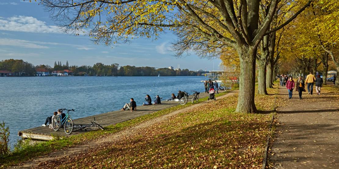 Menschen sitzen am Maschseeufer an einem Steg in der herbstlichen Sonne, Spaziergänger sind in der herbstlichen Allee am Seeufer zu sehen.